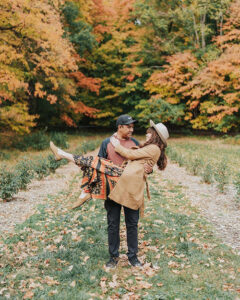 A man carries a woman in front of fall foliage.