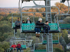 A mother and son sit on a chair lift with fall foliage in the background