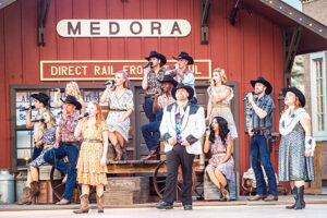 A group of young people in western wear sing into microphones in front of a red barn