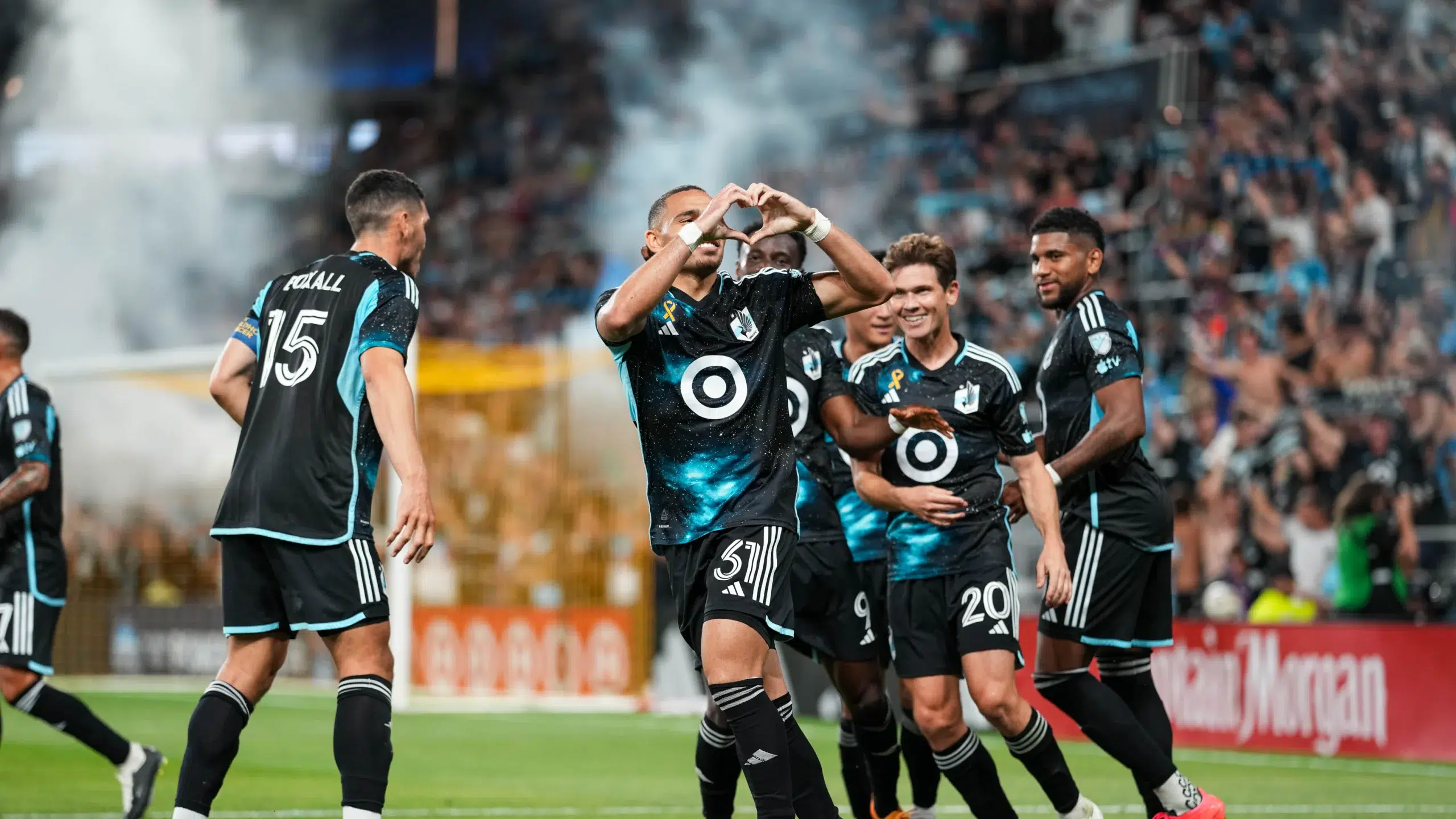 Multiple soccer players are on the field. One smiles to the crowd and makes a heart shape with his hands.