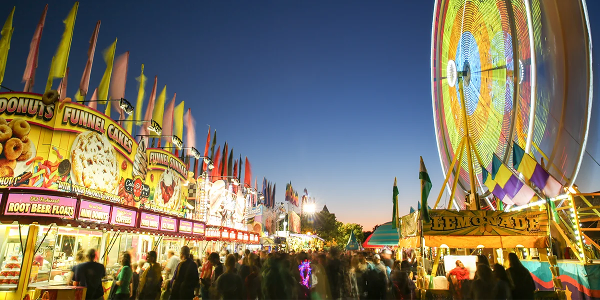 A carnival scene on the midway, with a moving wheel, signage for funnel cakes, and crowds of people