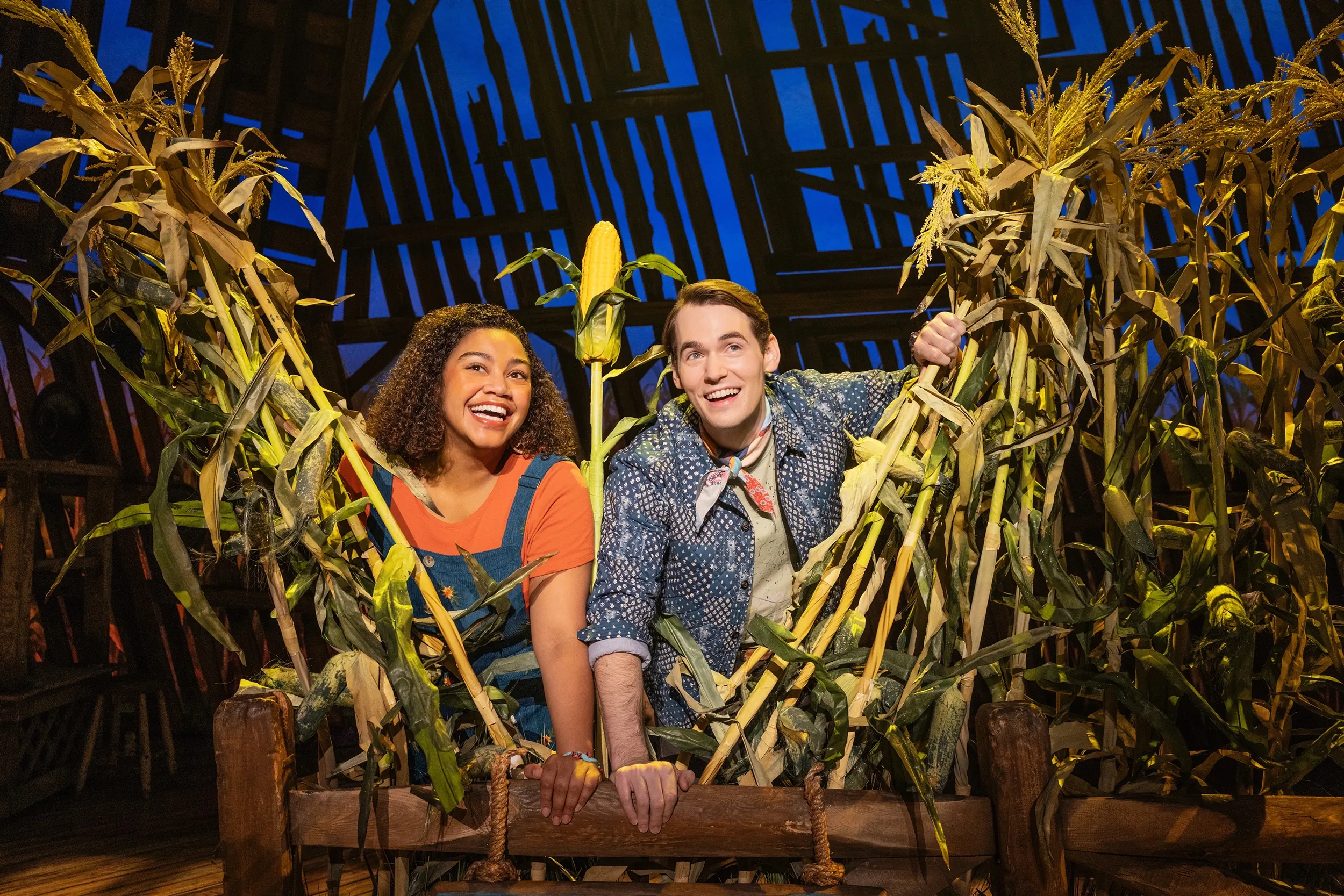 A smiling couple peeks out from behind tall corn