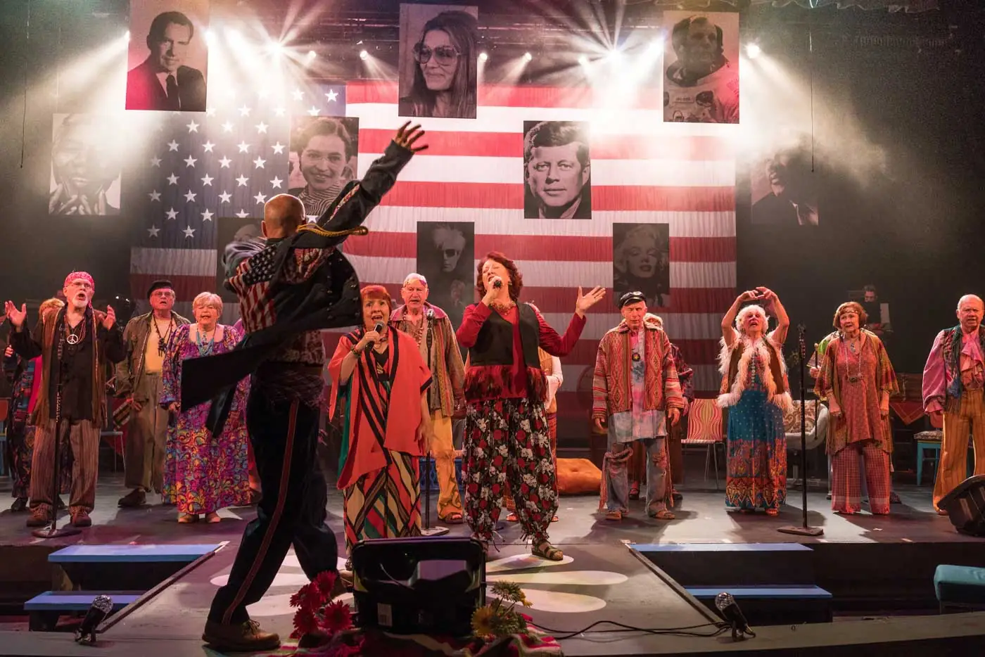 A group of seniors dressed in clothing from the 1960s perform in front of an American flag and photos of famous people from the era